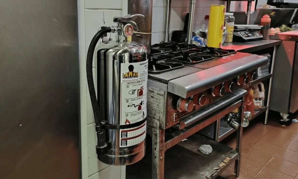 Extintor tipo K de acero inoxidable con etiqueta de servicio LAMAR, instalado en la pared de una cocina comercial junto a la estufa