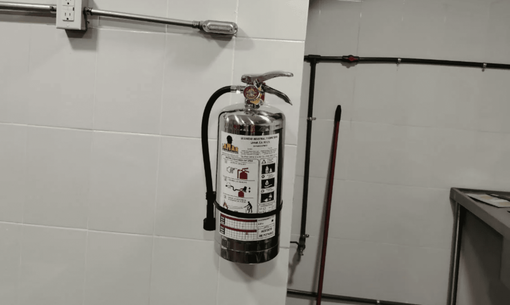 Extintor tipo K de acero inoxidable con etiqueta de servicio LAMAR, instalado en una columna de cocina industrial